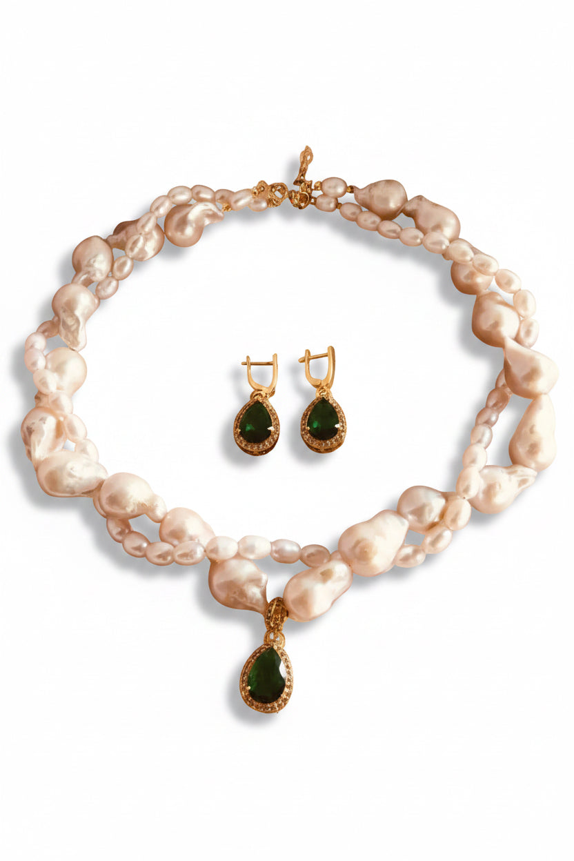 A pearl necklace and matching drop earrings displayed on a soft blush pink surface. The necklace features irregular white pearls with gold-toned links and a teardrop-shaped dark green stone pendant.