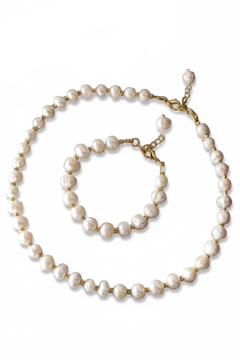 A pearl necklace and matching pearl bracelet with gold-tone chain links, arranged on a light marble surface with a burlap fabric and purple fabric prop in the background.