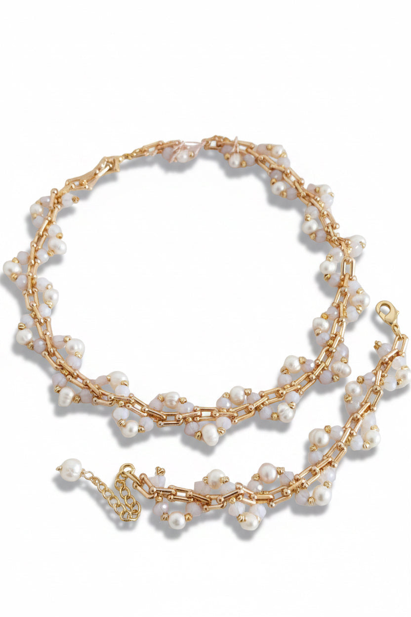 A gold chain necklace decorated with small white pearls, arranged in a loop on a light fabric surface with pink dried flowers and pale fluffy decor in the background.