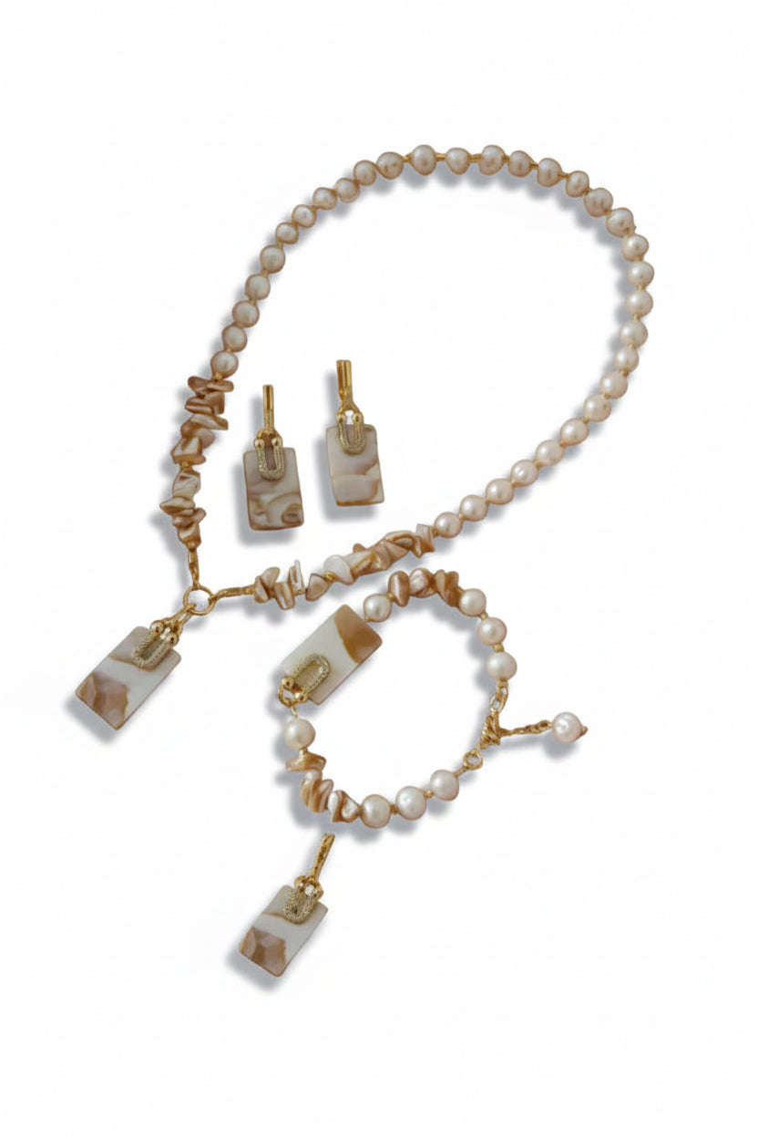 A jewelry set featuring a pearl and shell necklace, matching drop earrings, and a bracelet with gold-tone hardware and rectangular marbled stone pendants, arranged on a round blush pink tray with decorative dried flowers and white feathers.