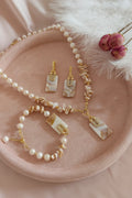 A jewelry set featuring a pearl and shell necklace, matching drop earrings, and a bracelet with gold-tone hardware and rectangular marbled stone pendants, arranged on a round blush pink tray with decorative dried flowers and white feathers.