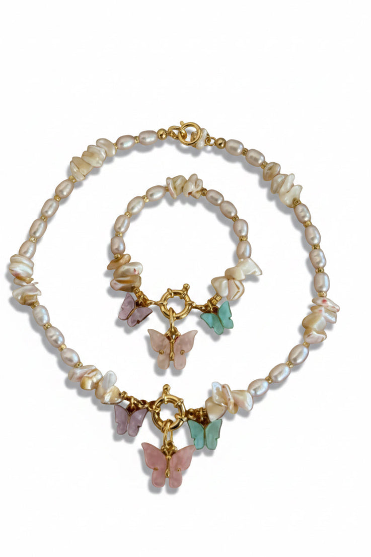 A matching jewelry set featuring a beaded necklace and bracelet with irregular white pearls, gold-tone hardware, and pastel butterfly charms in pink, purple, and mint green, displayed on a soft blush pink surface with white feathers at the top.