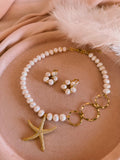 A pearl and gold jewelry set featuring a necklace with a starfish pendant, matching flower-shaped earrings, and gold rings, arranged on a soft beige fabric background with white feathers.