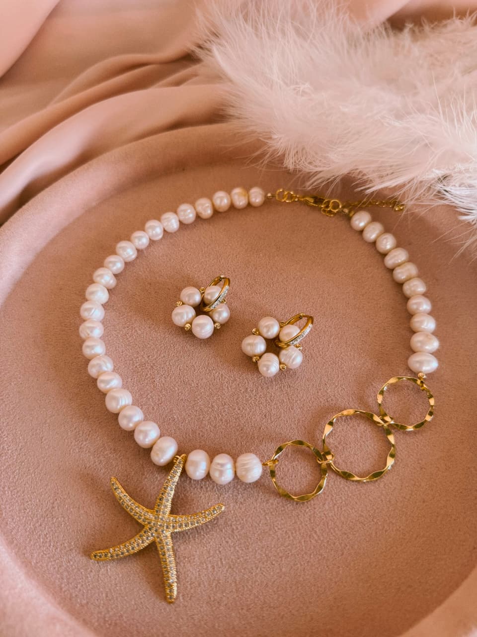 A pearl and gold jewelry set featuring a necklace with a starfish pendant, matching flower-shaped earrings, and gold rings, arranged on a soft beige fabric background with white feathers.