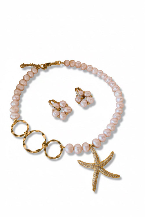 A pearl and gold jewelry set featuring a necklace with a starfish pendant, matching flower-shaped earrings, and gold rings, arranged on a soft beige fabric background with white feathers.